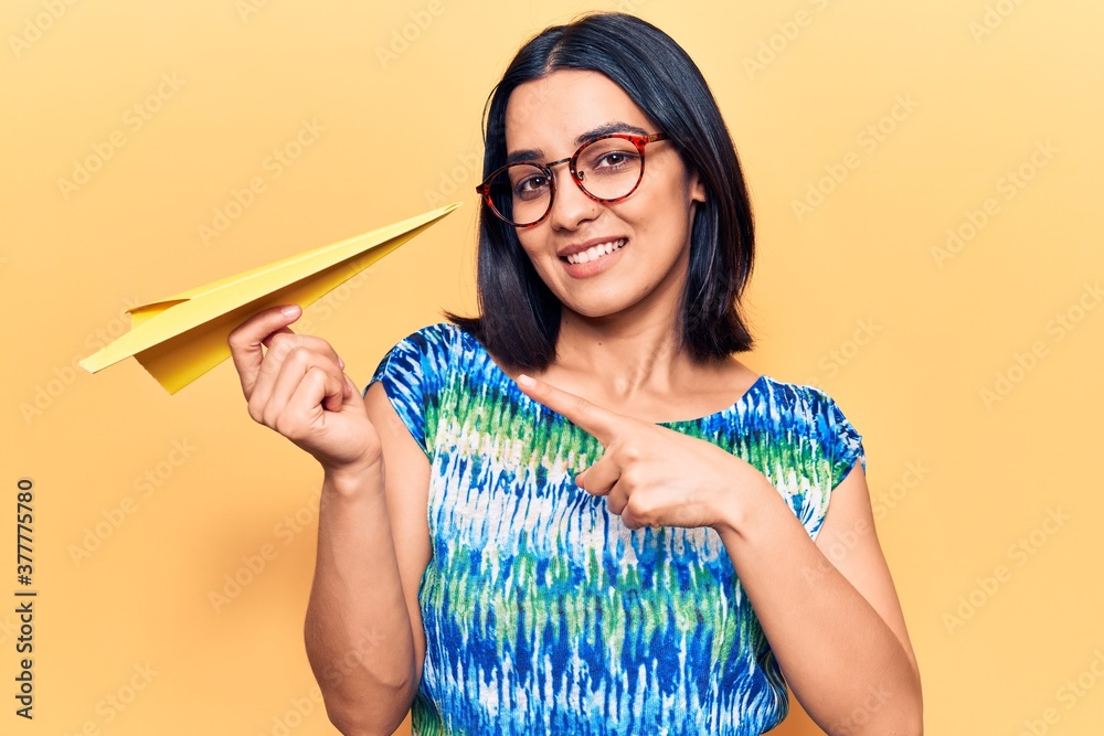 Young beautiful latin woman holding paper airplane smiling happy pointing with hand and finger Young beautiful latin woman holding paper airplane smiling happy pointing with hand and finger