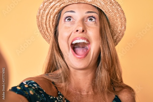 Middle age hispanic woman wearing summer hat taking a selfie angry and mad screaming frustrated and furious, shouting with anger looking up.
