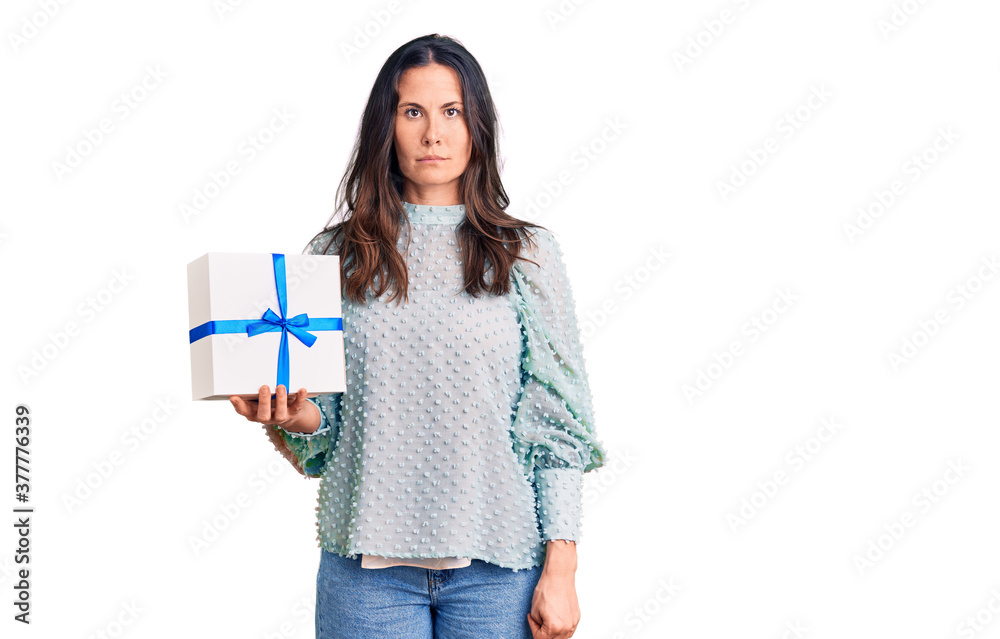 Young beautiful brunette woman holding birthday gift thinking attitude and sober expression looking self confident Young beautiful brunette woman holding birthday gift thinking attitude and sober expression looking self confident