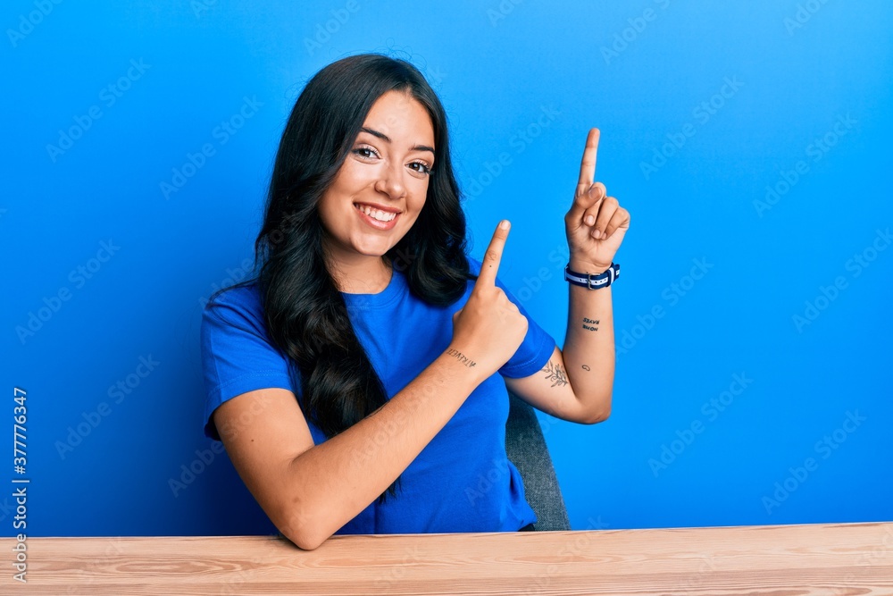 Beautiful brunette young woman wearing casual clothes sitting on the table smiling and looking at the camera pointing with two hands and fingers to the side. Beautiful brunette young woman wearing casual clothes sitting on the table smiling and looking at the camera pointing with two hands and fingers to the side.