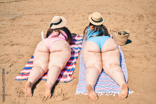 Backview of two plus size overweight sisters twins women relaxing lying on a towel at the beach on summer holidays