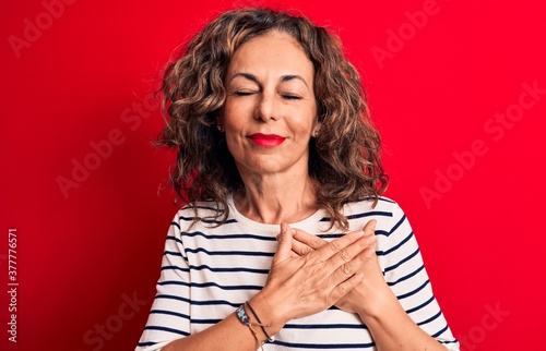 Middle age beautiful brunette woman wearing striped t-shirt standing over red background smiling with hands on chest, eyes closed with grateful gesture on face. Health concept.