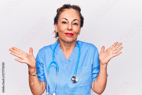 Fotografija Middle age brunette nurse woman wearing uniform and stethoscope over white background clueless and confused with open arms, no idea and doubtful face
