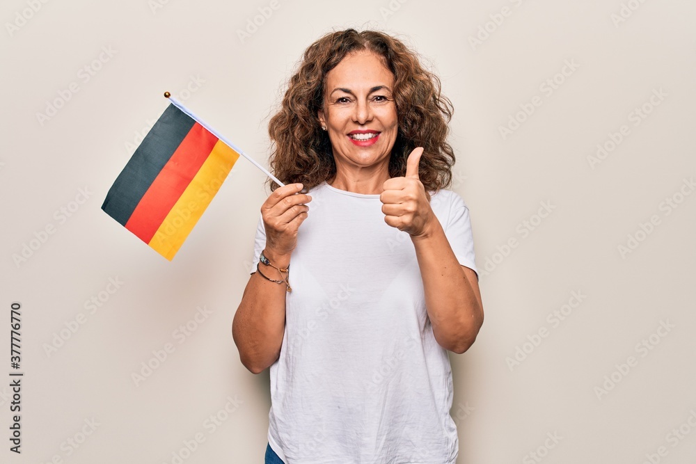 Middle age beautiful patriotic woman holding german flag over isolated white background smiling happy and positive, thumb up doing excellent and approval sign Middle age beautiful patriotic woman holding german flag over isolated white background smiling happy and positive, thumb up doing excellent and approval sign