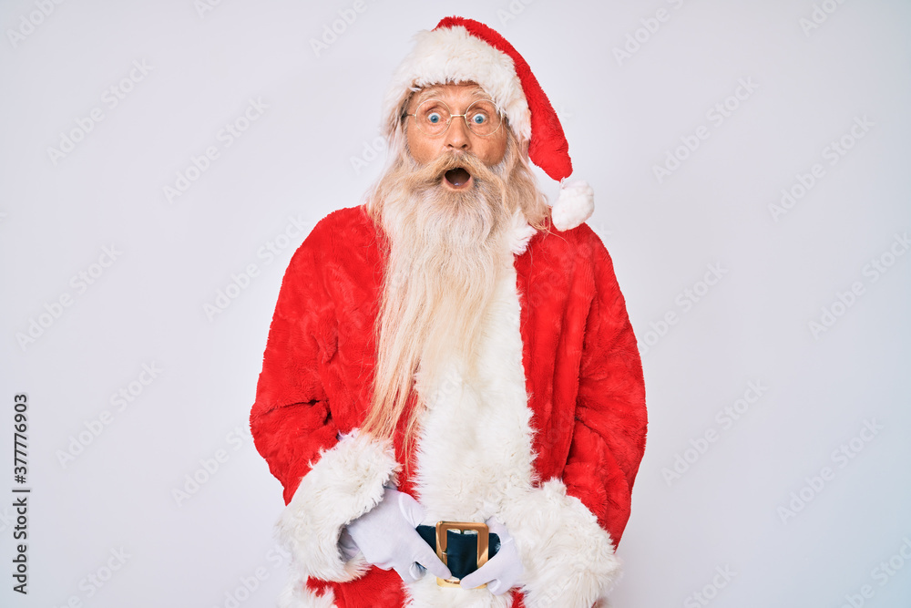 Old senior man with grey hair and long beard wearing santa claus costume with suspenders afraid and shocked with surprise expression, fear and excited face.
