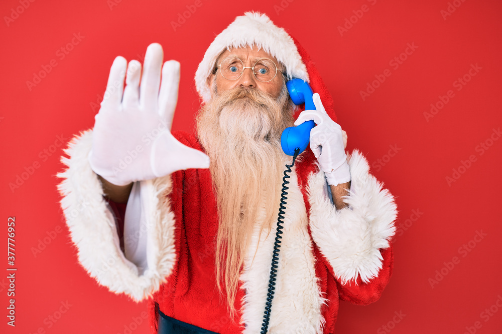 Old senior man wearing santa claus costume using vintage telephone with open hand doing stop sign with serious and confident expression, defense gesture Old senior man wearing santa claus costume using vintage telephone with open hand doing stop sign with serious and confident expression, defense gesture