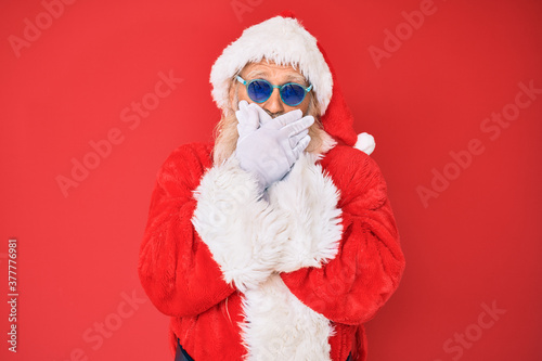 Old senior man wearing santa claus costume and sunglasses shocked covering mouth with hands for mistake. secret concept.