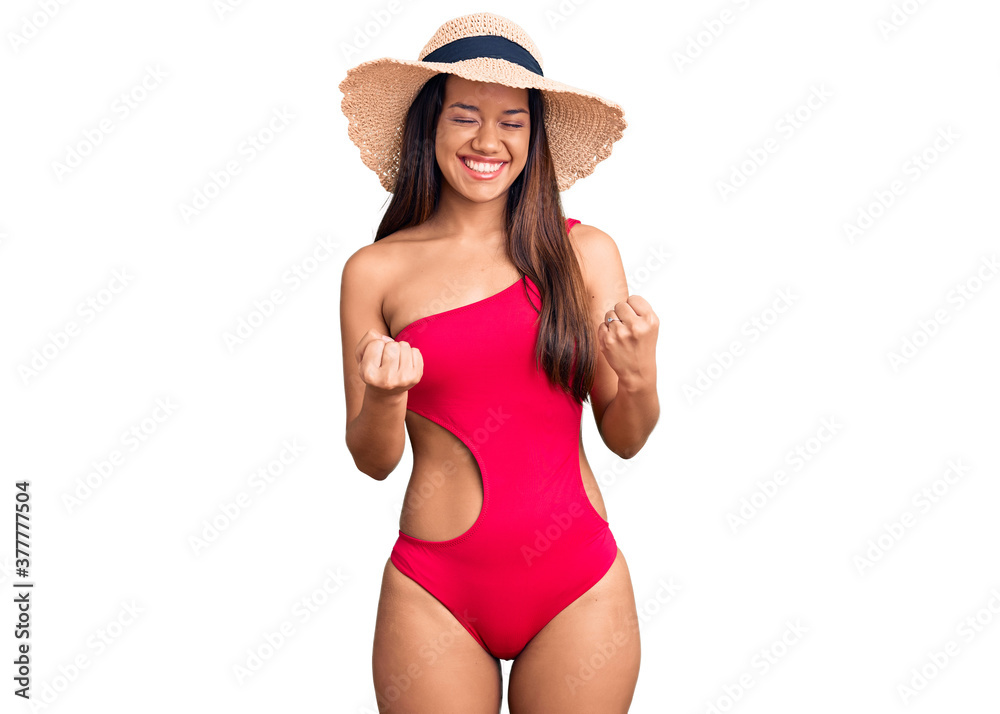 Young beautiful latin girl wearing swimwear and summer hat excited for success with arms raised and eyes closed celebrating victory smiling. winner concept. Young beautiful latin girl wearing swimwear and summer hat excited for success with arms raised and eyes closed celebrating victory smiling. winner concept.