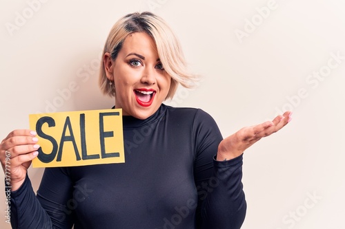 Young beautiful blonde plus size woman holding sale banner over isolated white background celebrating achievement with happy smile and winner expression with raised hand