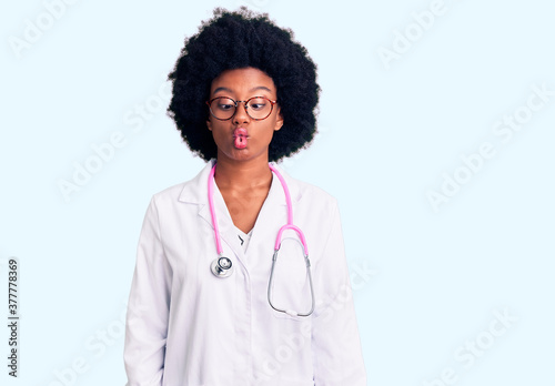 Young african american woman wearing doctor coat and stethoscope making fish face with lips, crazy and comical gesture. funny expression.