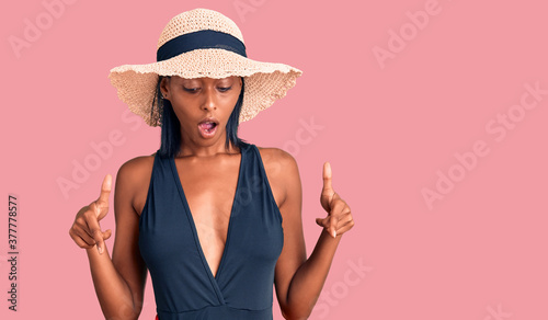 Young african american woman wearing swimsuit and summer hat pointing down with fingers showing advertisement, surprised face and open mouth