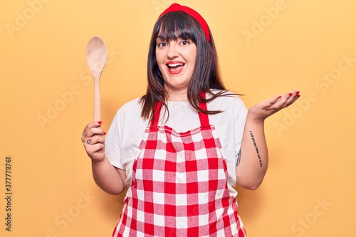 Young plus size woman wearing apron holding wooden spoon celebrating achievement with happy smile and winner expression with raised hand