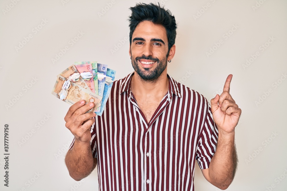Handsome hispanic man with beard holding canadian dollars smiling happy pointing with hand and finger to the side Handsome hispanic man with beard holding canadian dollars smiling happy pointing with hand and finger to the side