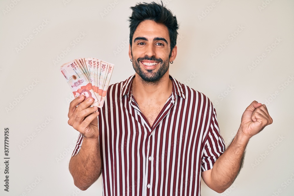 Handsome hispanic man with beard holding colombian pesos screaming proud, celebrating victory and success very excited with raised arm Handsome hispanic man with beard holding colombian pesos screaming proud, celebrating victory and success very excited with raised arm