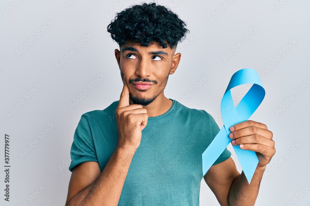 Young arab handsome man holding blue ribbon serious face thinking about question with hand on chin, thoughtful about confusing idea Young arab handsome man holding blue ribbon serious face thinking about question with hand on chin, thoughtful about confusing idea