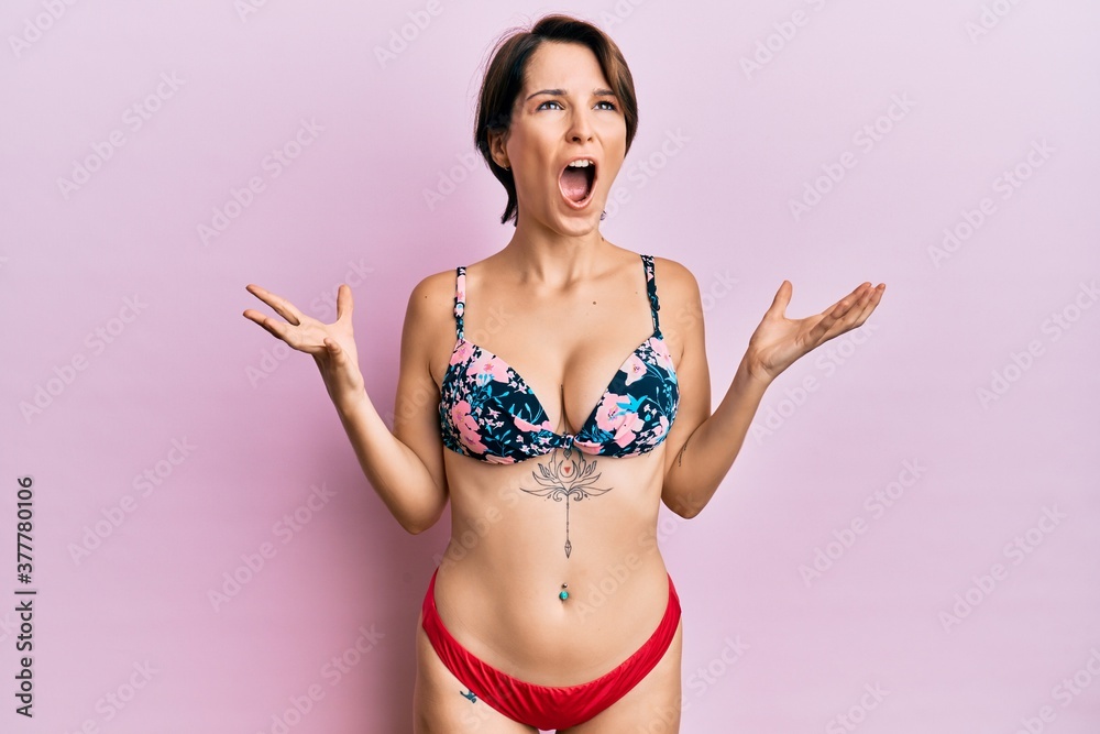 Young brunette woman with short hair wearing bikini showing tattoos crazy and mad shouting and yelling with aggressive expression and arms raised. frustration concept. Young brunette woman with short hair wearing bikini showing tattoos crazy and mad shouting and yelling with aggressive expression and arms raised. frustration concept.