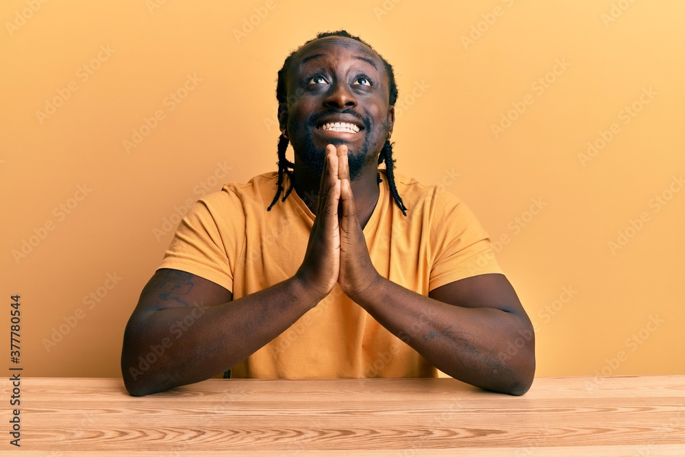 Handsome young black man wearing casual clothes sitting on the table begging and praying with hands together with hope expression on face very emotional and worried. begging. Handsome young black man wearing casual clothes sitting on the table begging and praying with hands together with hope expression on face very emotional and worried. begging.