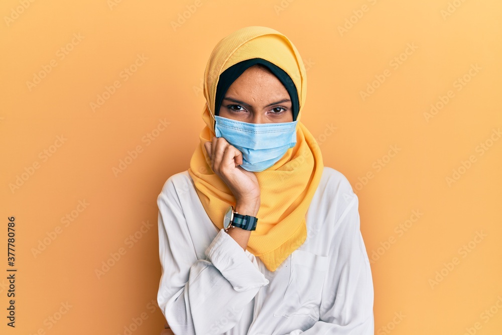 Young brunette arab woman wearing traditional islamic hijab and medical mask thinking looking tired and bored with depression problems with crossed arms. Young brunette arab woman wearing traditional islamic hijab and medical mask thinking looking tired and bored with depression problems with crossed arms.