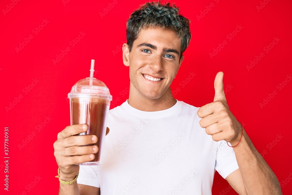 Young handsome man holding glass of smoothie smiling happy and positive, thumb up doing excellent and approval sign Young handsome man holding glass of smoothie smiling happy and positive, thumb up doing excellent and approval sign