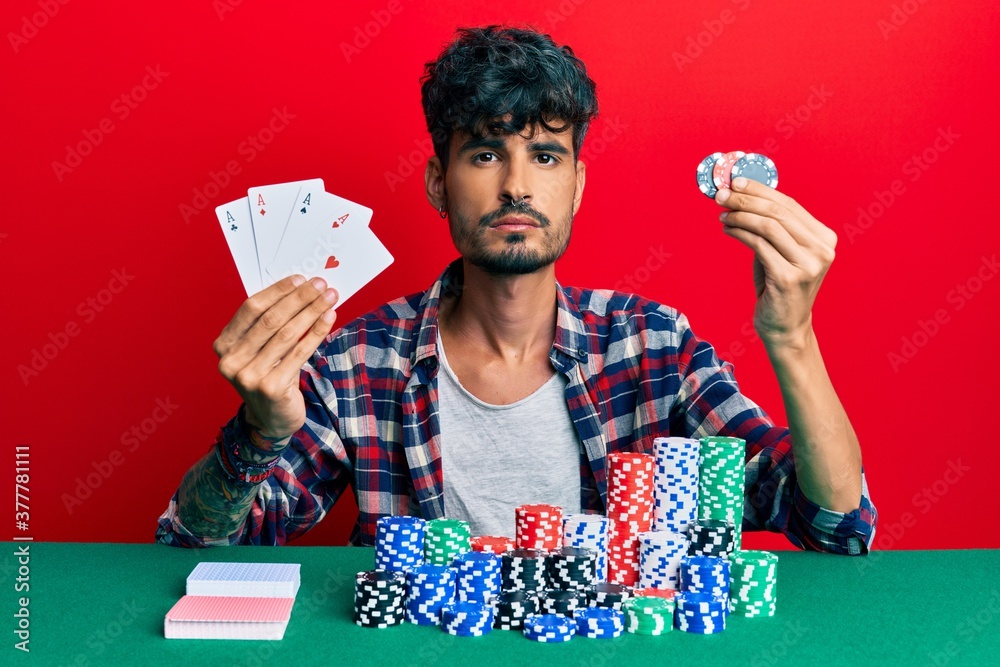 Young hispanic man playing poker holding cards and casino chips relaxed with serious expression on face. simple and natural looking at the camera. Young hispanic man playing poker holding cards and casino chips relaxed with serious expression on face. simple and natural looking at the camera.