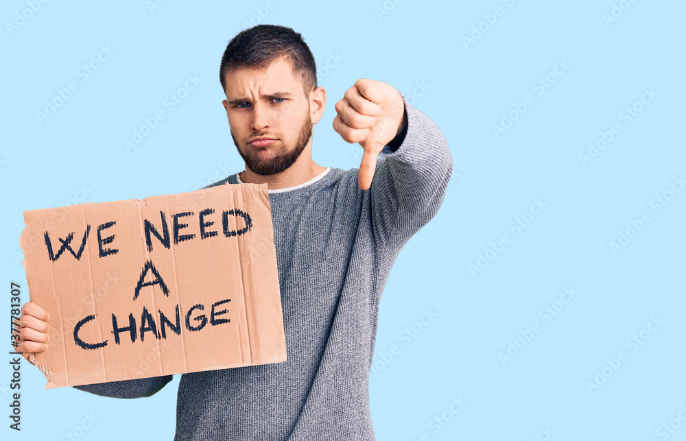 Young handsome man holding we need a change banner with angry face, negative sign showing dislike with thumbs down, rejection concept Young handsome man holding we need a change banner with angry face, negative sign showing dislike with thumbs down, rejection concept