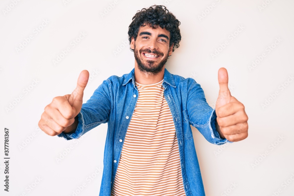 Handsome young man with curly hair and bear wearing casual denim jacket approving doing positive gesture with hand, thumbs up smiling and happy for success. winner gesture. Handsome young man with curly hair and bear wearing casual denim jacket approving doing positive gesture with hand, thumbs up smiling and happy for success. winner gesture.