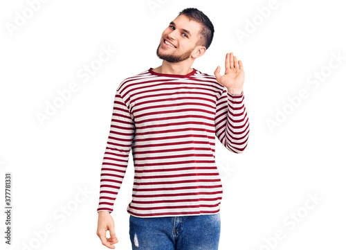 Young handsome man wearing striped sweater waiving saying hello happy and smiling, friendly welcome gesture