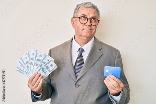 Senior grey-haired man wearing business suit holding credit car and dollars smiling looking to the side and staring away thinking.