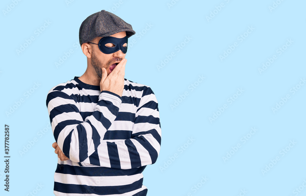 Young handsome man wearing burglar mask bored yawning tired covering mouth with hand. restless and sleepiness.