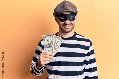 Young handsome bald man wearing burglar mask stealing dollars banknotes looking positive and happy standing and smiling with a confident smile showing teeth photo