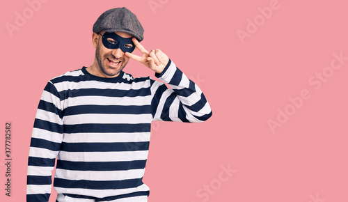 Young handsome man wearing burglar mask doing peace symbol with fingers over face, smiling cheerful showing victory