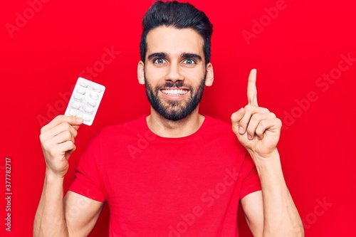 Young handsome man with beard holding pills smiling with an idea or question pointing finger with happy face, number one photo