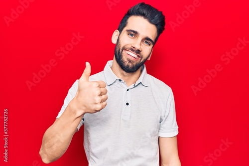 Young handsome man with beard wearing casual polo doing happy thumbs up gesture with hand. approving expression looking at the camera showing success.
