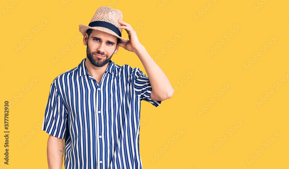 Young handsome man with beard wearing summer hat and striped shirt confuse and wonder about question. uncertain with doubt, thinking with hand on head. pensive concept.