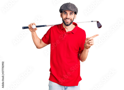 Young handsome man with beard playing golf holding club and ball smiling happy pointing with hand and finger to the side