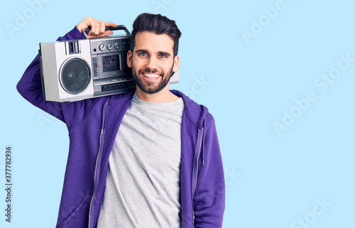 Young handsome man with beard listening to music using vintage boombox looking positive and happy standing and smiling with a confident smile showing teeth