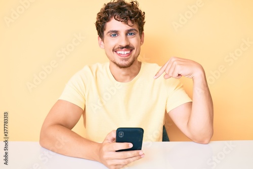 Young caucasian man with curly hair using smartphone sitting on the table pointing finger to one self smiling happy and proud