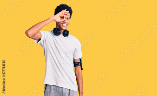 Young african american man wearing gym clothes and using headphones smiling happy doing ok sign with hand on eye looking through fingers