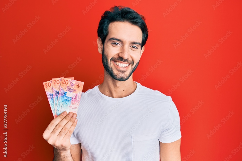 Young hispanic man holding swiss franc banknotes looking positive and happy standing and smiling with a confident smile showing teeth Young hispanic man holding swiss franc banknotes looking positive and happy standing and smiling with a confident smile showing teeth