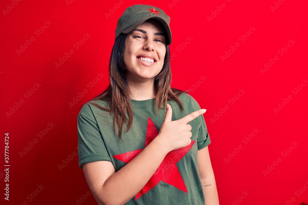 Young beautiful brunette woman wearing cap and t-shirt with red star communist symbol smiling cheerful pointing with hand and finger up to the side Young beautiful brunette woman wearing cap and t-shirt with red star communist symbol smiling cheerful pointing with hand and finger up to the side