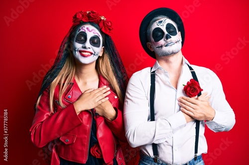 Couple wearing day of the dead costume over red smiling with hands on chest with closed eyes and grateful gesture on face. health concept.