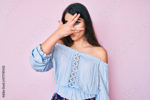 Brunette teenager girl wearing casual clothes peeking in shock covering face and eyes with hand, looking through fingers afraid