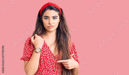 Brunette teenager girl wearing summer dress in hurry pointing to watch time, impatience, looking at the camera with relaxed expression © Krakenimages.com