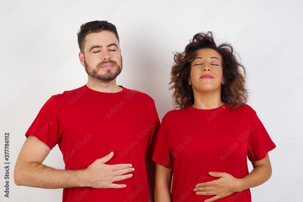 Young beautiful couple wearing red t-shirt on white background touches tummy, smiles gently, eating and satisfaction concept. Young beautiful couple wearing red t-shirt on white background touches tummy, smiles gently, eating and satisfaction concept.