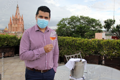 Casual latin man with protection mask, tasting rosé sparkling wine in crystal goblet
in outdoor terrace with view of the cathedral in San Miguel de Allende Guanajuato Mexico, new normal photo