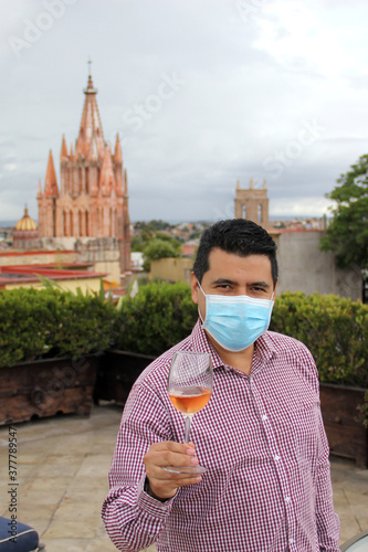 Casual latin man with protection mask, tasting rosé sparkling wine in crystal goblet
in outdoor terrace with view of the cathedral in San Miguel de Allende Guanajuato Mexico, new normal photo