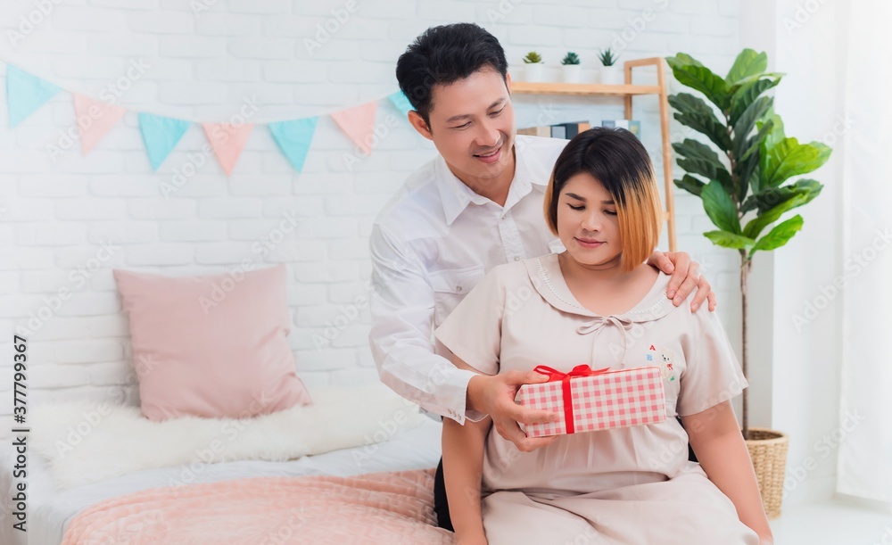 Asian husband holds a gift box to a young wife who is pregnant in bedroom at home.Concept of maintaining health during a happy pregnancy.