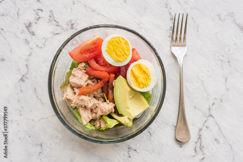 Top down closeup of healthy salad in glass bowl with tuna, egg, avocado, tomato and lettucs on marble effect background with fork (selective focus)