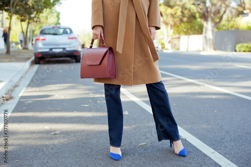 Fashionable young woman wearing beige wool coat, blue jeans and blue high heel shoes. She is holding burgundy handbag in hand.