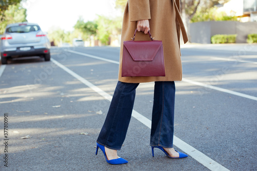 Fashionable young woman wearing beige wool coat, blue jeans and blue high heel shoes. She is holding burgundy handbag in hand.